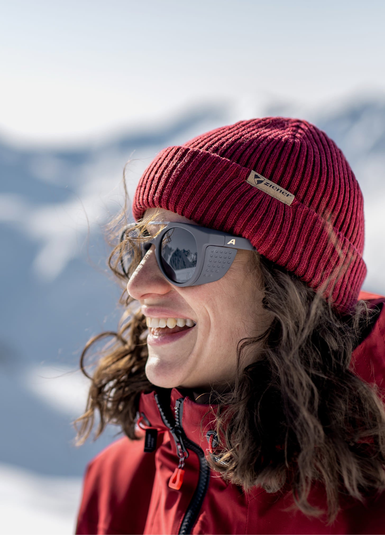 Laughing woman in a red beanie and sunglasses in front of snowy mountains