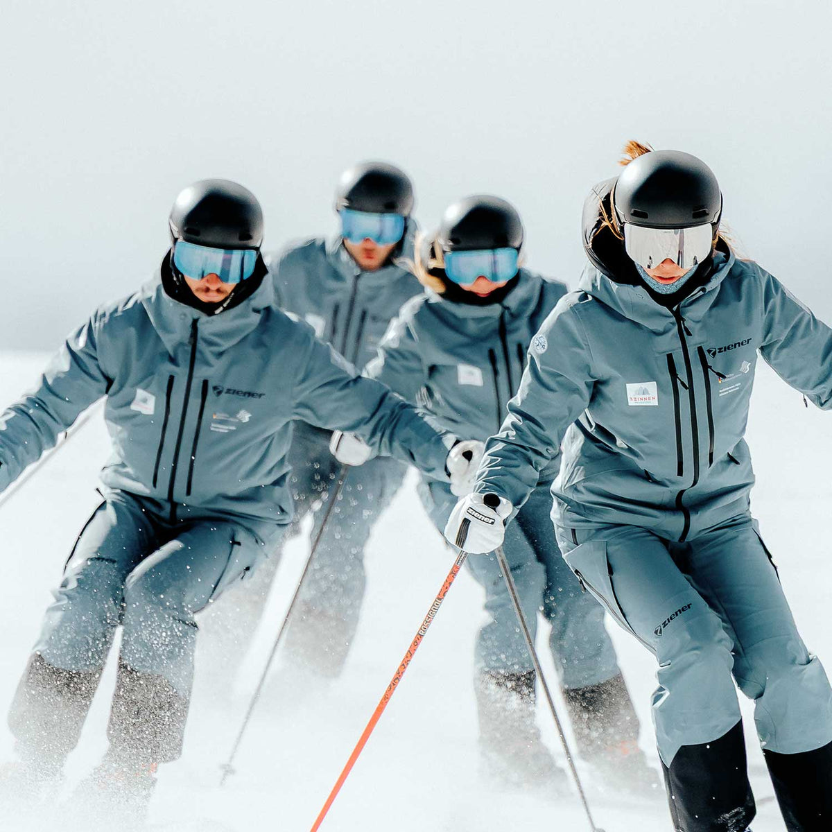 Vier Skilehrer in grauer Teamkleidung mit Helm und Skibrille fahren in Formation