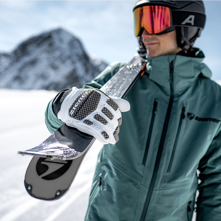 Skifahrer in grüner Jacke trägt Ski über der Schulter, im Vordergrund ein weißer ZIENER Rennhandschuh