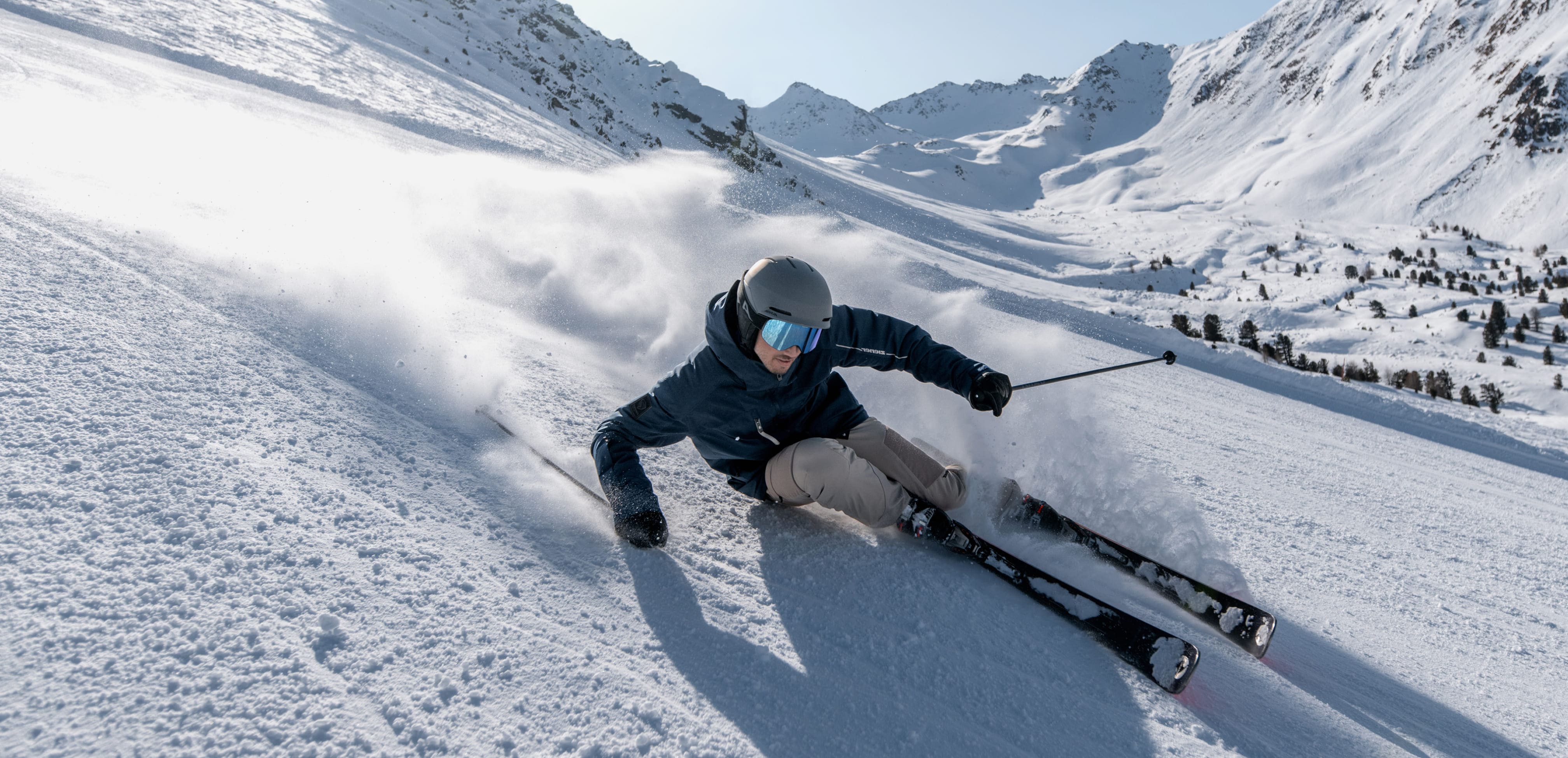 Skier carving through spraying snow in the sunshine with a mountain panorama behind