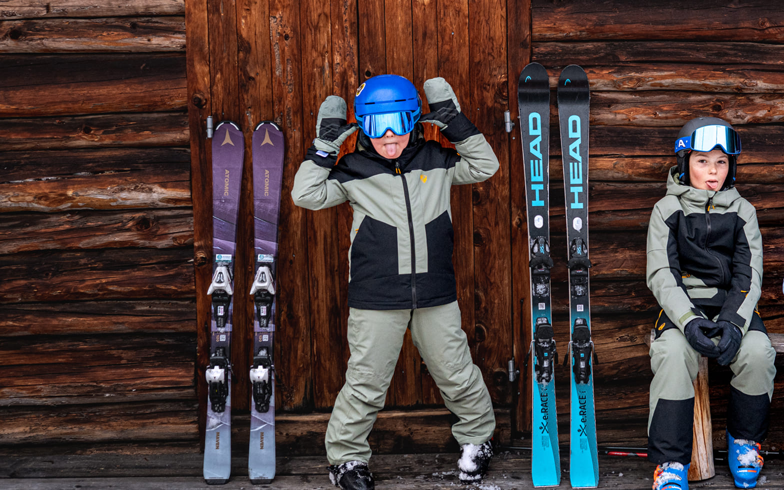 Zwei Kinder in Skibekleidung mit Helm und Skibrille vor einer Holzhütte, daneben ihre Ski