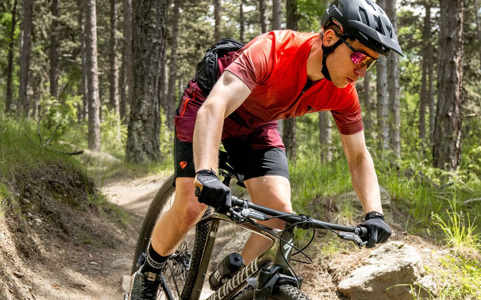 Mountainbiker in rotem Trikot fährt über einen Trail im Wald